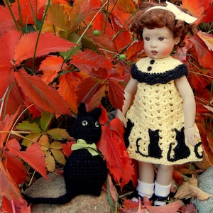 Può includere: Una bambola con i capelli castani e un vestito di uncinetto giallo con gatti neri. La bambola è in piedi su un cumulo di foglie rosse e arancioni con un giocattolo di gatto di uncinetto nero al suo fianco.