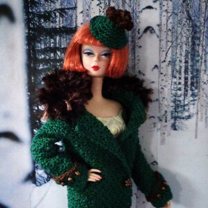 May include: A doll wearing a green crocheted coat with brown fur trim, a brown crocheted hat with a flower accent, and a yellow dress.