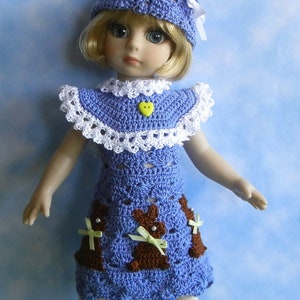 May include: A doll wearing a blue crocheted dress with white trim and brown bunny appliques. The dress has a yellow heart button. The doll is wearing a blue crocheted hat with a white ribbon and white tights. The doll is standing on a green surface.