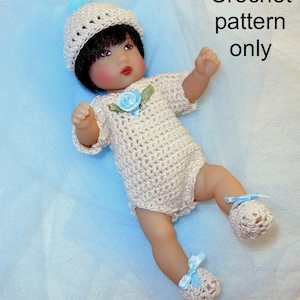 Puede incluir: Patrón de ganchillo solamente. Un conjunto de bebé de ganchillo con un gorro blanco y zapatitos azules. El conjunto es beige y tiene un adorno de flor azul.