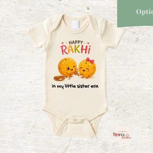 May include: A cream-colored short-sleeve baby bodysuit featuring a graphic of two smiling orange characters. The text reads HAPPY RAKHI in colorful letters and in my little sister era in black script below the characters.