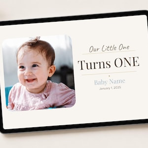 May include: A digital tablet displays a photo of a smiling baby in a pink shirt. Text on the screen reads "Our Little One Turns ONE" with space for a baby's name and the date January 1, 2025. A white stylus rests on the right.