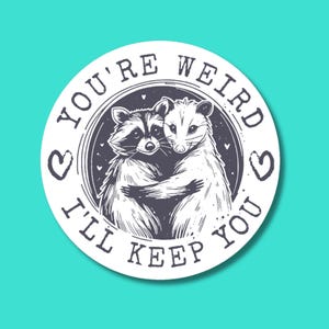 May include: A white circle sticker with black text that reads "You're Weird I'll Keep You" with a heart symbol on either side of the text.  The sticker features a black and white illustration of a raccoon and a possum hugging.