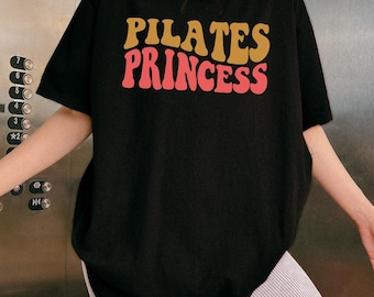 May include: Black t-shirt with the text "PILATES PRINCESS" in a retro font. The text is in shades of gold and red. The shirt is a casual, oversized fit.