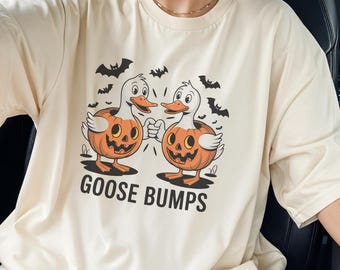 May include: Cream-colored t-shirt with a Halloween-themed graphic. The design features two cartoon ducks with pumpkin heads bumping fists, surrounded by bats. The text "GOOSE BUMPS" is printed below the ducks.