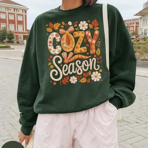 Puede incluir: Sudadera verde bosque con las palabras "COZY Season" en un diseño de temática otoñal. El diseño incluye elementos florales y a cuadros en naranja, amarillo y blanco. La sudadera se combina con pantalones cortos a rayas rosas y blancas.