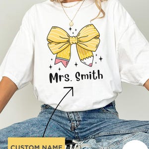 May include: A white t-shirt featuring a graphic of a yellow bow made from pencils with pink erasers and black outlines. Below the bow, the text reads Mrs. Smith in a black sans-serif font. Small black star icons are scattered around the bow design.