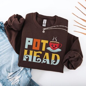 May include: A brown sweatshirt with the words "POT HEAD" in colorful block letters and a cartoon coffee cup graphic. A silver necklace is draped across the sweatshirt. The sweatshirt is paired with ripped blue jeans.