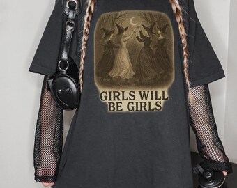 May include: Black oversized t-shirt featuring a vintage-style illustration of four witches dancing under a crescent moon. The text "GIRLS WILL BE GIRLS" is printed below the image in a serif font. The shirt has short sleeves and a distressed neckline.