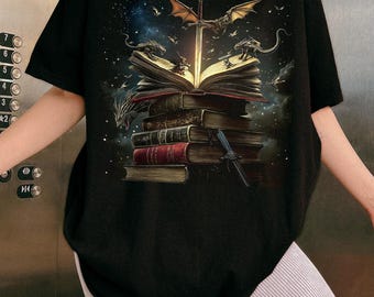 May include: Black t-shirt featuring a fantasy illustration. A stack of books is topped with an open book, a sword, and dragons. The design includes a starry sky and flying creatures, creating a magical theme.