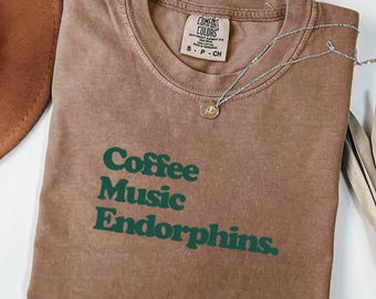 May include: Espresso-colored t-shirt with the words "Coffee Music Endorphins" in green block letters. A silver necklace with a small pendant rests on the shirt. The shirt is made by Comfort Colors.
