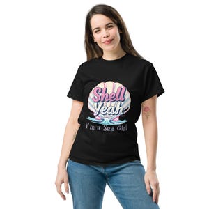 Può includere: T-shirt nera con grafica a conchiglia. La grafica presenta le parole "Shell Yeah" in rosa e blu, con la frase "I'm a Sea Girl" sotto. La conchiglia è nelle tonalità del rosa, bianco e blu.