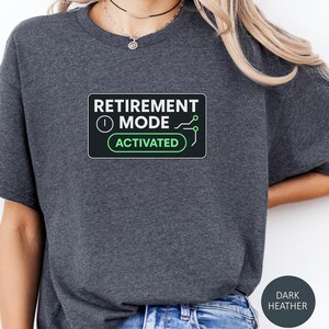 May include: Dark heather gray t-shirt with a graphic that reads "RETIREMENT MODE ACTIVATED" in white and green. The design is in a black rectangle with rounded corners. The shirt is a casual style.
