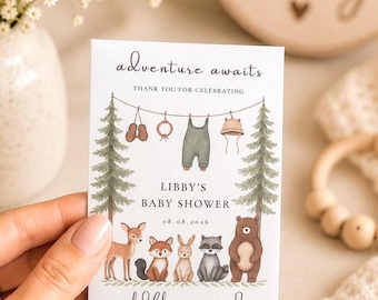 May include: A baby shower invitation card with the text "adventure awaits" and "Libby's Baby Shower". The card features illustrations of woodland animals, baby clothes, and pine trees. The words "wildflower seeds" are also printed on the card.