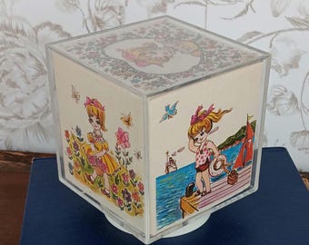 May include: A clear acrylic cube music box featuring illustrated panels. One side shows a child in a yellow dress walking through flowers, while another side depicts a child in a polka dot swimsuit standing on a wooden pier by the blue sea.