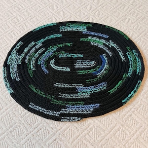 May include: A round, black crocheted rug with a spiral design. The rug features accents of blue, green, and white in a repeating pattern. The rug is made of a thick yarn and has a textured appearance. The rug is suitable for indoor use.