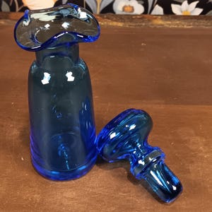 May include: A cobalt blue glass bottle with a unique, flared top and a separate stopper. The bottle has a cylindrical body and a rounded base. The stopper is a matching blue and has a decorative, swirling design.
