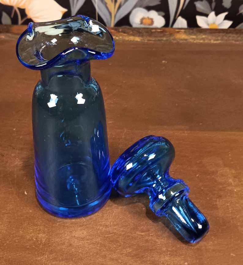 May include: A cobalt blue glass bottle with a unique, flared top and a separate stopper. The bottle has a cylindrical body and a rounded base. The stopper is a matching blue and has a decorative, swirling design.