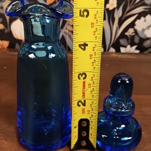 May include: A cobalt blue glass vase with a ruffled rim, next to a small, matching stoppered bottle. The vase is approximately 5 inches tall. The bottle is approximately 2 inches tall. Both are on a wooden surface.