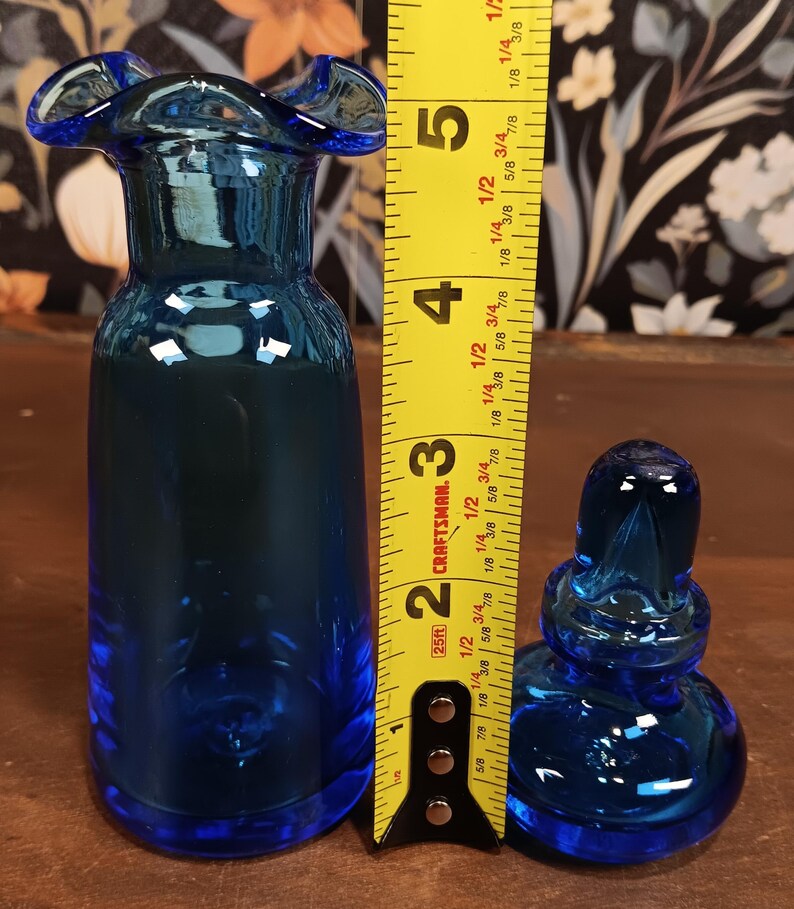 May include: A cobalt blue glass vase with a ruffled rim, next to a small, matching stoppered bottle. The vase is approximately 5 inches tall. The bottle is approximately 2 inches tall. Both are on a wooden surface.