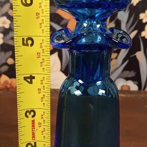 May include: A cobalt blue glass bottle with a bulbous top and flared accents. The bottle is approximately 6 inches tall. A yellow measuring tape is positioned next to the bottle.