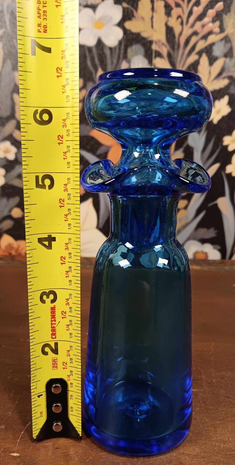 May include: A cobalt blue glass bottle with a bulbous top and flared accents. The bottle is approximately 6 inches tall. A yellow measuring tape is positioned next to the bottle.