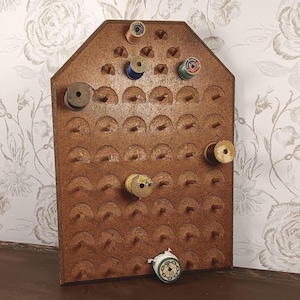 May include: A brown wooden thread rack with a pointed top and multiple rows of pegs. Several vintage spools of thread in various colors, including blue, green, and yellow, are placed on the pegs, while one white spool rests at the base.