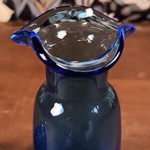 May include: A close-up of a cobalt blue glass vase with a flared rim. The vase has a rounded body and a wide, open top. White and light blue flecks are visible on the rim and inside the vase. The background is blurred.