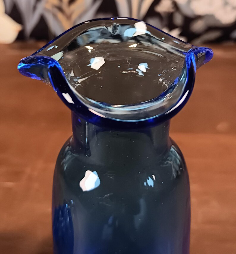 May include: A close-up of a cobalt blue glass vase with a flared rim. The vase has a rounded body and a wide, open top. White and light blue flecks are visible on the rim and inside the vase. The background is blurred.