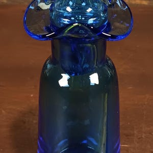 May include: A cobalt blue glass bottle with a unique, flower-like top. The bottle has a rounded base and a long neck. A yellow tape measure is in the foreground.