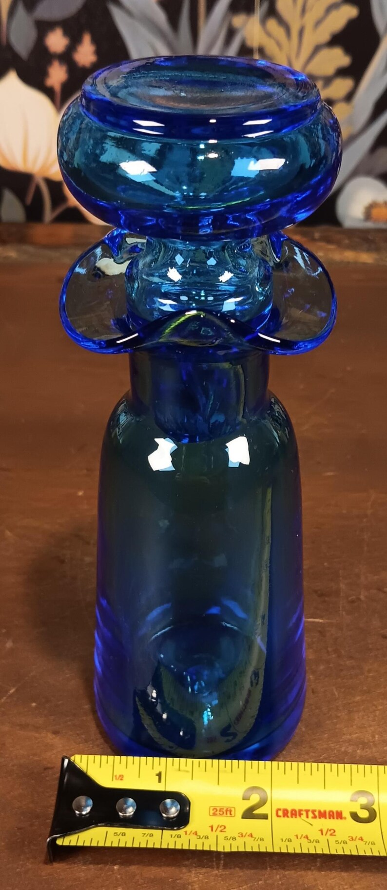 May include: A cobalt blue glass bottle with a unique, flower-like top. The bottle has a rounded base and a long neck. A yellow tape measure is in the foreground.