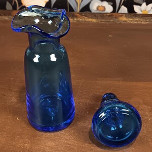 May include: A cobalt blue glass vase with a flared, ruffled rim, next to a matching stopper. The vase has a slender, elongated body. The stopper is rounded. Both pieces are on a brown surface.