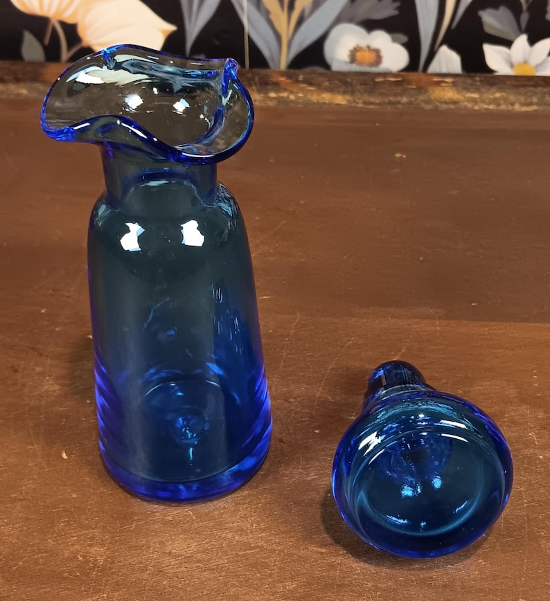 May include: A cobalt blue glass vase with a flared, ruffled rim, next to a matching stopper. The vase has a slender, elongated body. The stopper is rounded. Both pieces are on a brown surface.