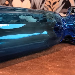May include: A blue glass bottle with a rounded base and a flared top. The bottle has a long, cylindrical body and a bulbous stopper. The glass has a glossy finish and reflects light. The bottle is lying on a brown surface.