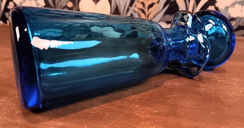 May include: A blue glass bottle with a rounded base and a flared top. The bottle has a long, cylindrical body and a bulbous stopper. The glass has a glossy finish and reflects light. The bottle is lying on a brown surface.