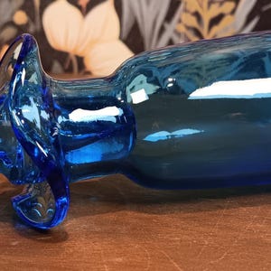 May include: A vibrant blue glass bottle with a unique, curved design. The bottle features a rounded base and a decorative handle. The glass has a glossy finish, reflecting light. The bottle is a decorative item.