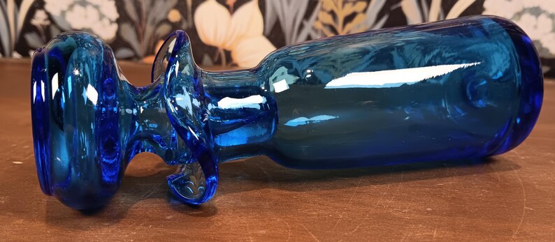 May include: A vibrant blue glass bottle with a unique, curved design. The bottle features a rounded base and a decorative handle. The glass has a glossy finish, reflecting light. The bottle is a decorative item.
