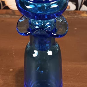 May include: A cobalt blue glass bottle with a unique design. The bottle features a bulbous top, a flared midsection, and a rounded base. The glass has a glossy finish, reflecting light. The bottle is likely a decorative item or a vase.