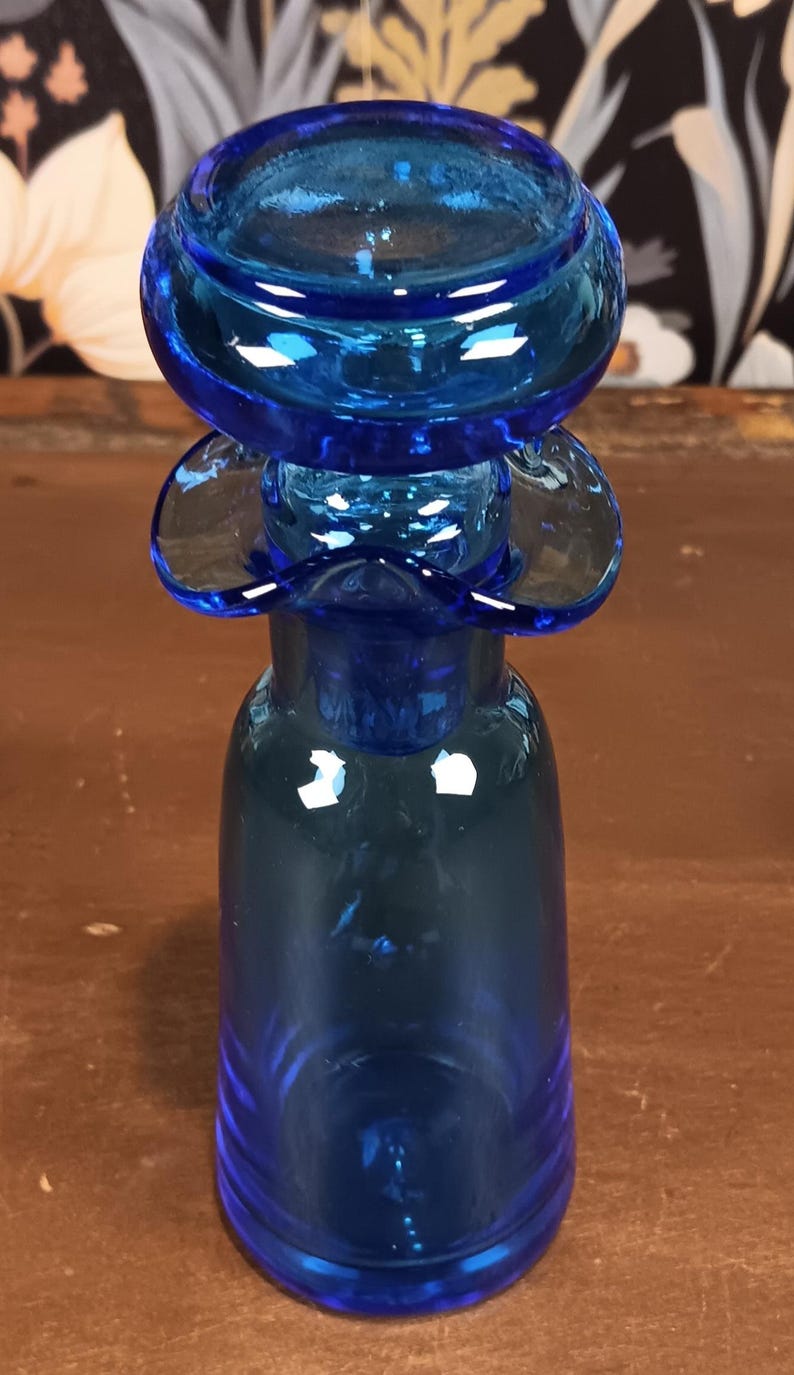May include: A cobalt blue glass bottle with a unique design. The bottle features a bulbous top, a flared midsection, and a rounded base. The glass has a glossy finish, reflecting light. The bottle is likely a decorative item or a vase.