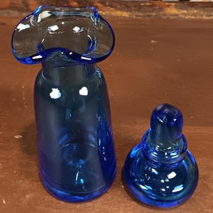 May include: A cobalt blue glass vase with a flared rim and a matching stopper. The vase has a rounded body and a ruffled top. The stopper is a rounded shape with a small knob. The glass is transparent and reflects light.