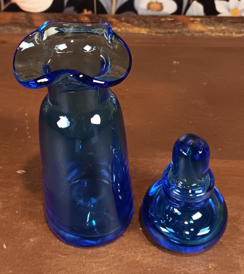 May include: A cobalt blue glass vase with a flared rim and a matching stopper. The vase has a rounded body and a ruffled top. The stopper is a rounded shape with a small knob. The glass is transparent and reflects light.
