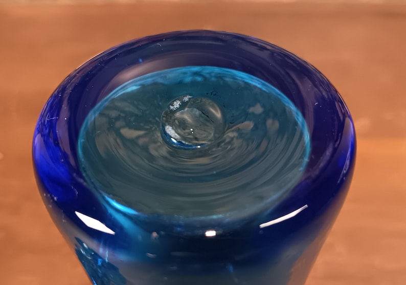 May include: A close-up view of a blue glass vase with a unique, swirling interior design. The vase features a deep blue exterior that transitions to a lighter turquoise hue inside, creating a captivating visual effect. The glass appears smooth and reflective.