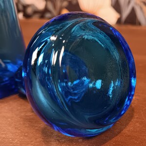 May include: A close-up of a round, blue glass object with a swirling pattern. The glass has a glossy finish and reflects light, creating highlights and shadows. The object appears to be a decorative piece or part of a larger glass item.
