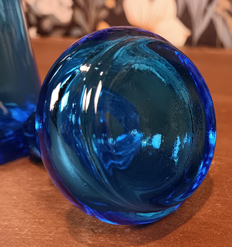 May include: A close-up of a round, blue glass object with a swirling pattern. The glass has a glossy finish and reflects light, creating highlights and shadows. The object appears to be a decorative piece or part of a larger glass item.