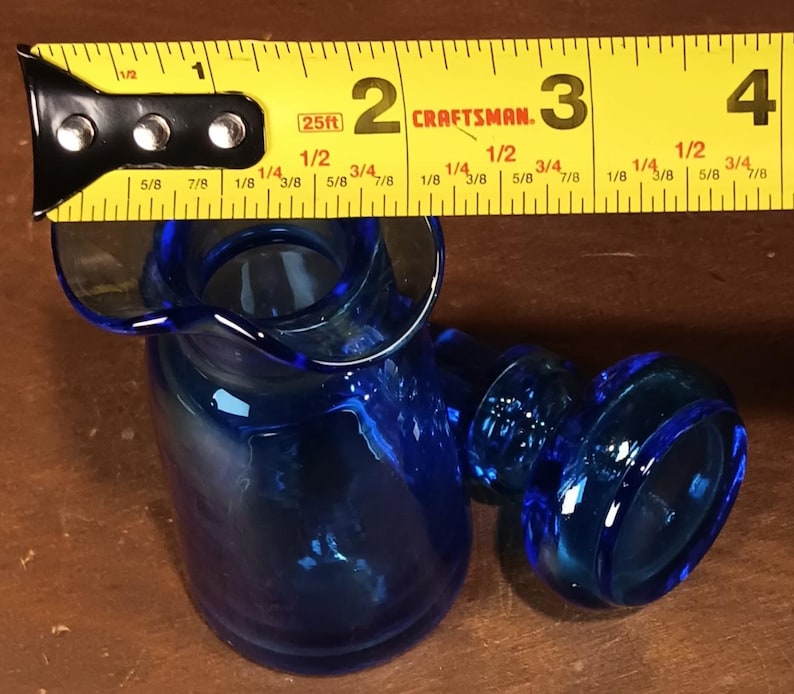 May include: A cobalt blue glass creamer with a flared spout and handle. The creamer is approximately 3 inches tall, as measured by a yellow Craftsman tape measure.