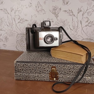 May include: A vintage Polaroid Minute Maker camera with a black strap rests on a patterned box and a tan book. The camera has a silver face with the Polaroid logo and the words "Minute Maker". The box has a black and white speckled design.