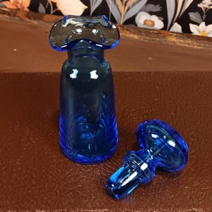 May include: A cobalt blue glass bottle with a matching stopper. The bottle has a flared top and a cylindrical body. The stopper is mushroom-shaped. The objects are on a brown surface. Decorative glassware.