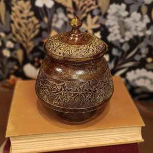 May include: A decorative, lidded jar with a detailed leaf pattern. The jar is brown with gold accents and sits atop a stack of books. The lid has a small, pointed finial. The jar's design suggests it could be used for storage or display.