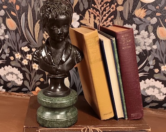 May include: A dark bronze bust of a young person sits atop a green marble base. Three antique books lean against each other, with titles visible. The background features a floral wallpaper design.
