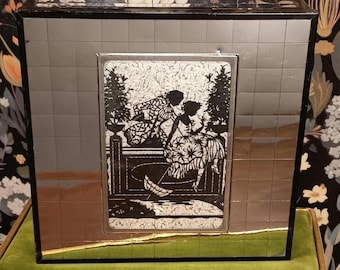 May include: A mirrored box with a black frame and a central panel featuring a black and white illustration of two figures in a garden setting. The box has a grid-like pattern on its mirrored surfaces.